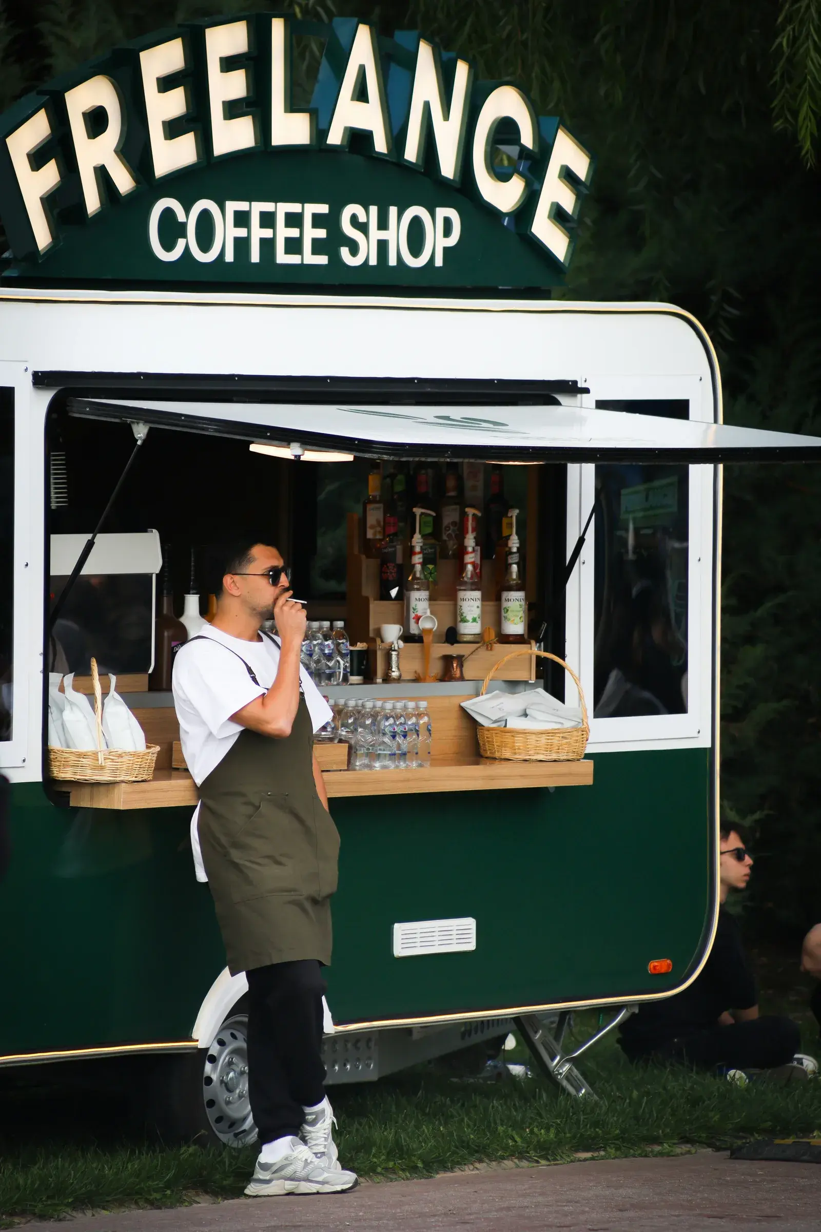 A barista in an apron standing outside a green coffee trailer signed Freelance Coffee Shop